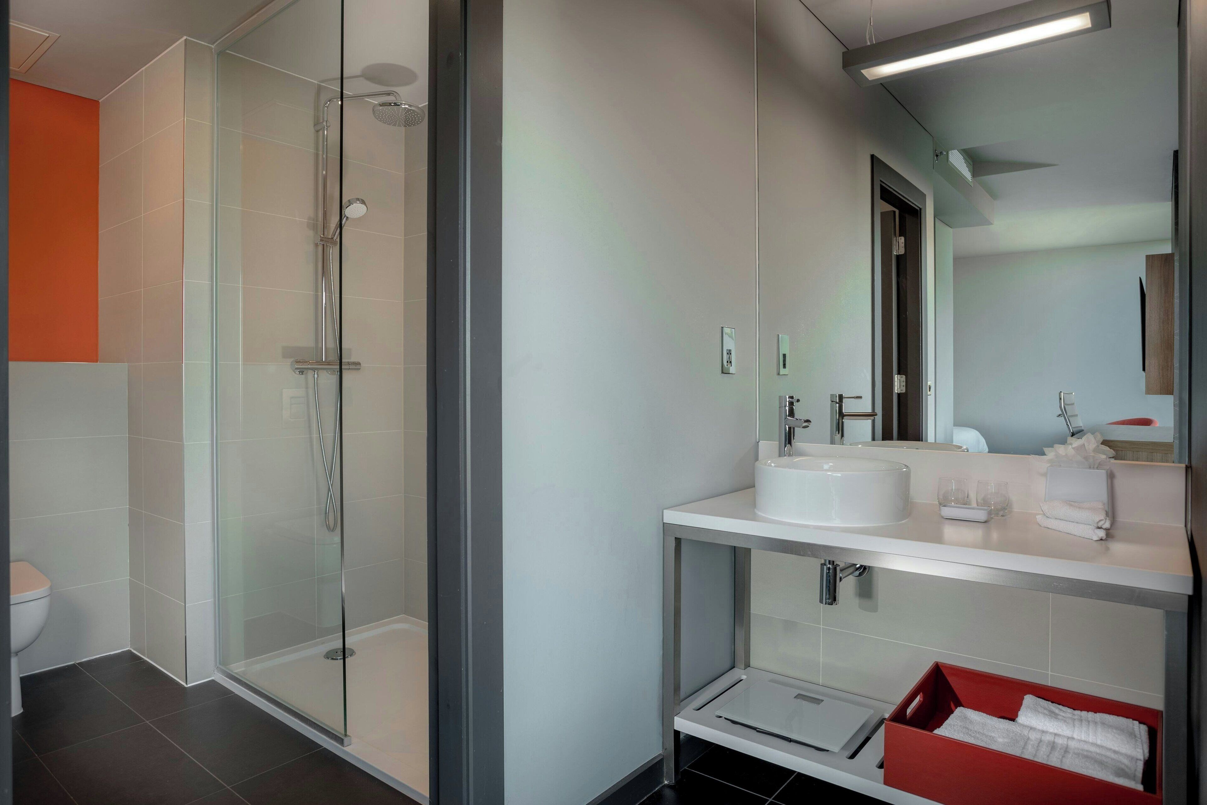 Un baño moderno con ducha envuelta en vidrio, un lavamanos blanco y un caja roja para guardar objetos.
