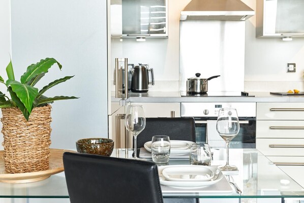 A modern kitchen with a glass dining table, white cabinets, stainless steel appliances, and a potted plant. - Vizion Serviced Apartments S (Milton Keynes)