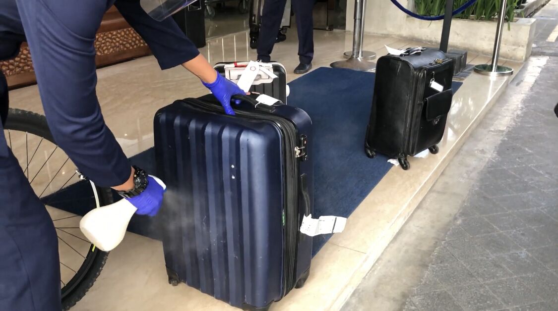 Una persona sostiene una maleta con una etiqueta, de pie junto a una zona alfombrada de color azul, posiblemente en un hotel o aeropuerto.