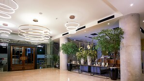 A spacious lobby with a modern design, featuring a large reception desk, multiple hanging light fixtures, and several large potted plants.