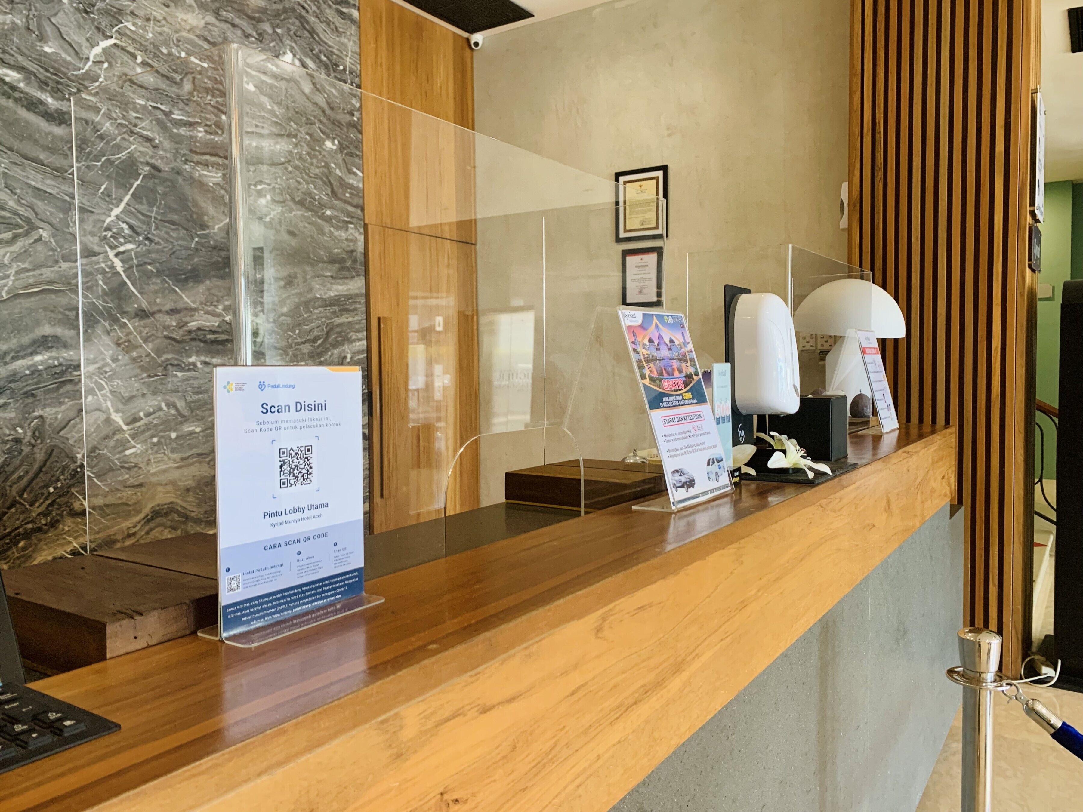 A hotel reception desk with a marble wall, wooden paneling, and a clear glass partition. There are signs for room service and a QR code for check-in.