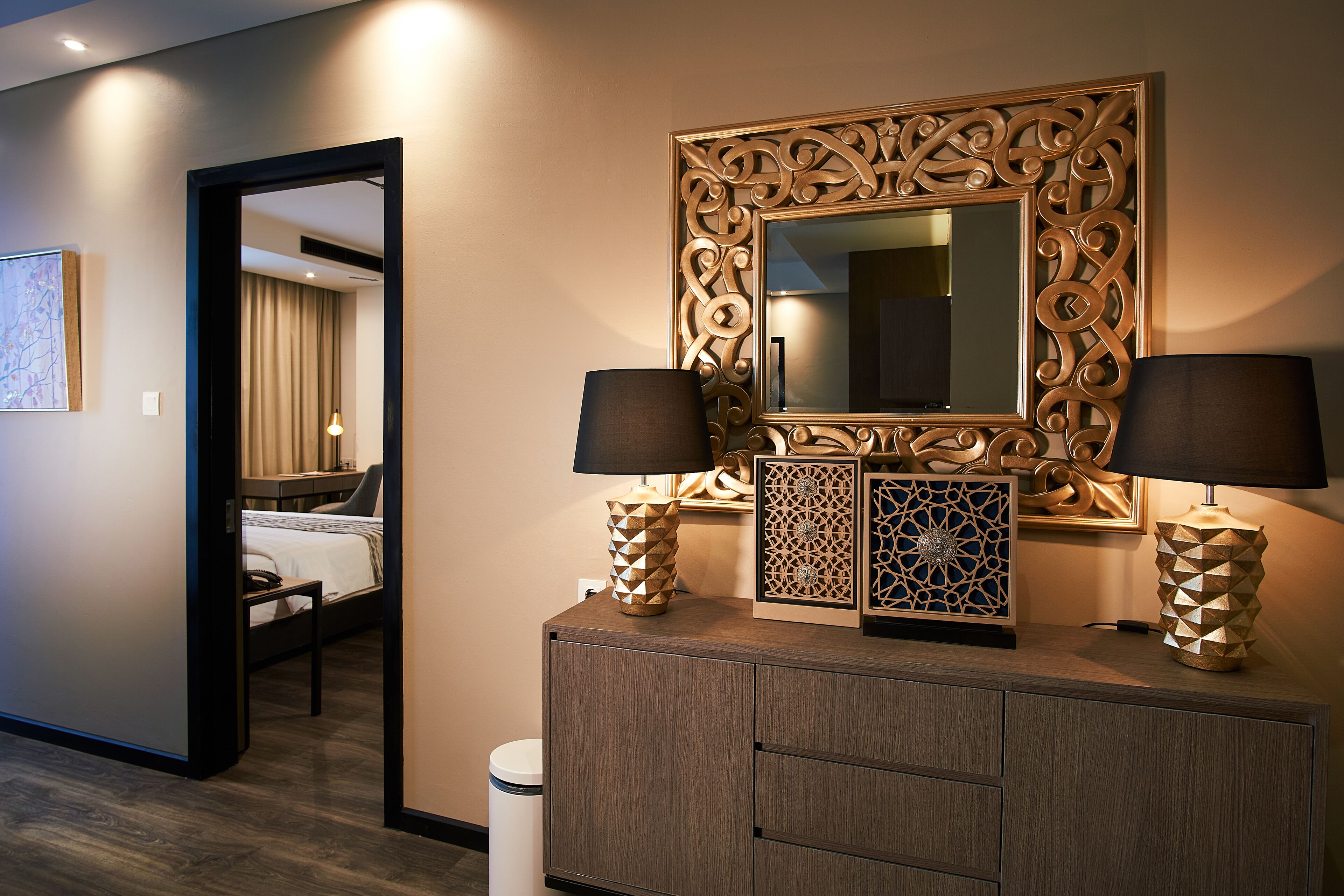A hotel room with a bed, a dresser with decorative items, a mirror with an ornate frame, and a door leading to another room.