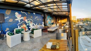 A rooftop lounge with wicker furniture, a mural with coffee-related text, and a view of the cityscape.