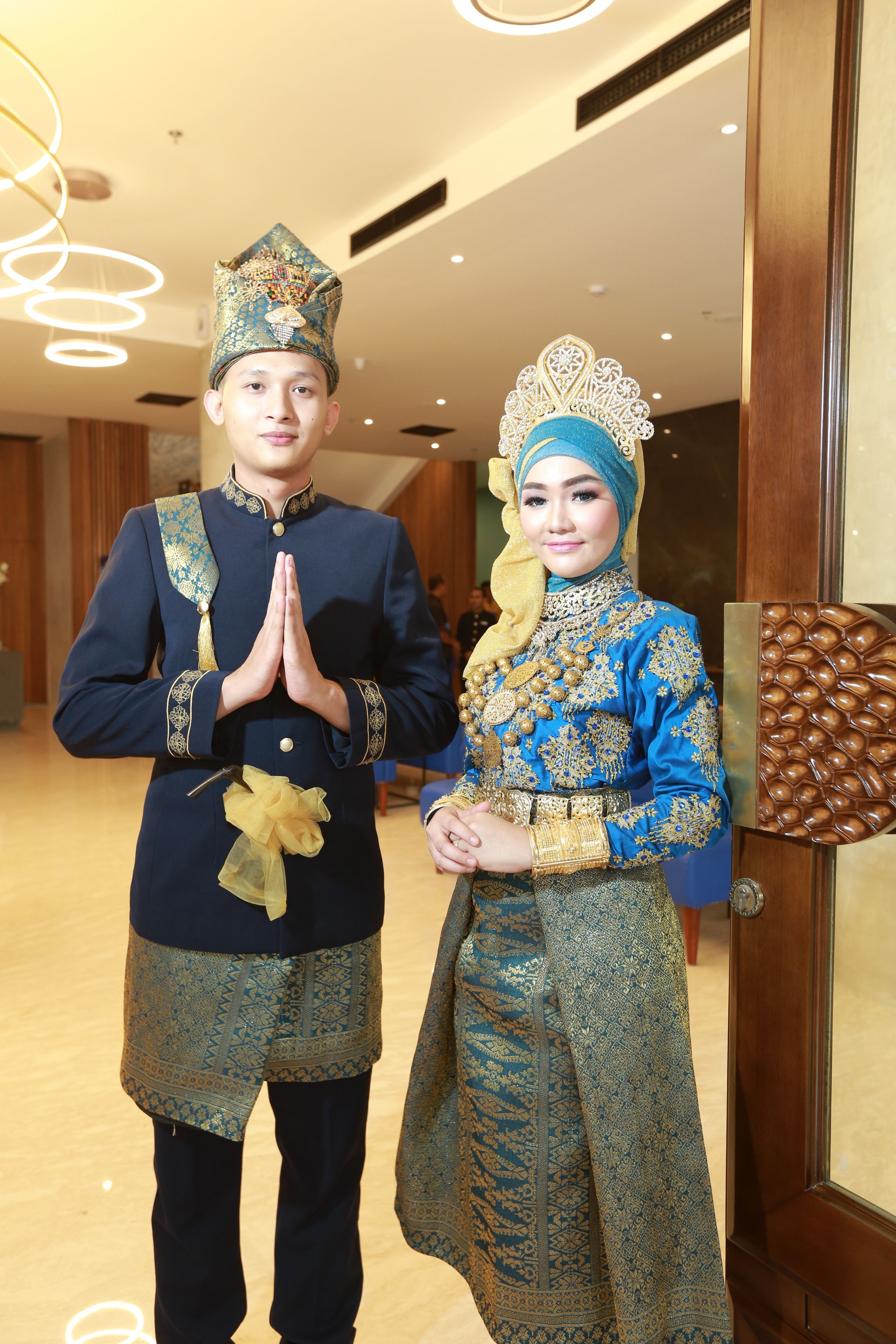 Un homme et une femme, vêtus de costumes traditionnels, se tiennent dans un couloir dont l’intérieur présente un design moderne, avec notamment une porte en bois et un luminaire.
