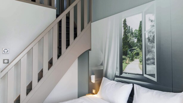 Quarto com cama, mesa de cabeceira, luminária na parede, janela com vista para árvores e escada.