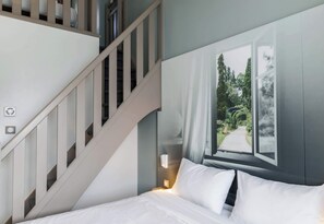A bedroom with a bed, a nightstand, a wall-mounted lamp, a window with a view of trees, and a staircase. - B&B HOTEL Nancy Frouard (1) (Frouard)