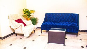 A room with a blue sofa, a white armchair with a red pillow, a small table, and a potted plant. - Hotel Quo Vadis (Abuja)