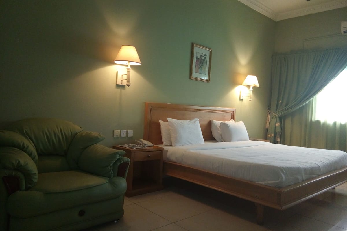 A hotel room with a bed, a green armchair, bedside lamps, and a painting on the wall.