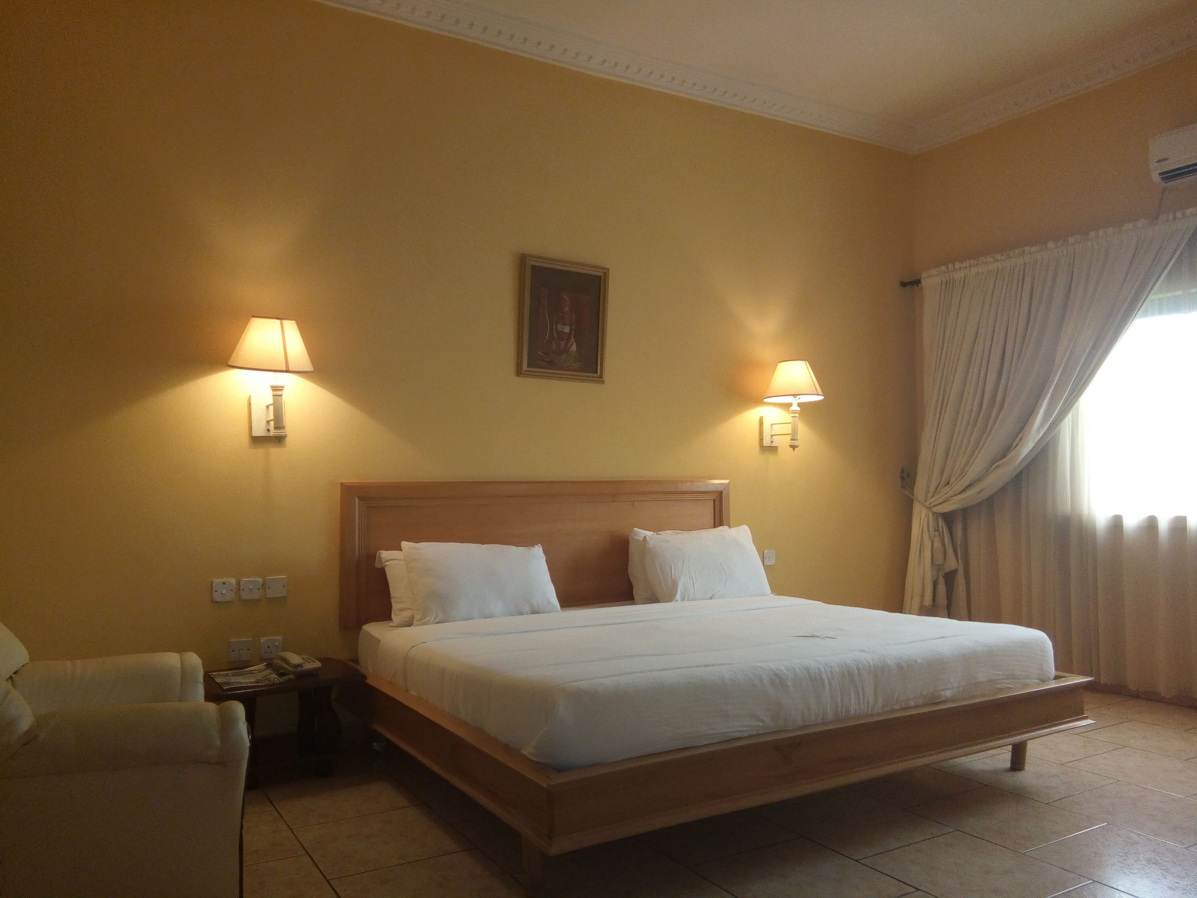 A hotel room with a large bed, two bedside lamps, a painting on the wall, and a window with curtains.