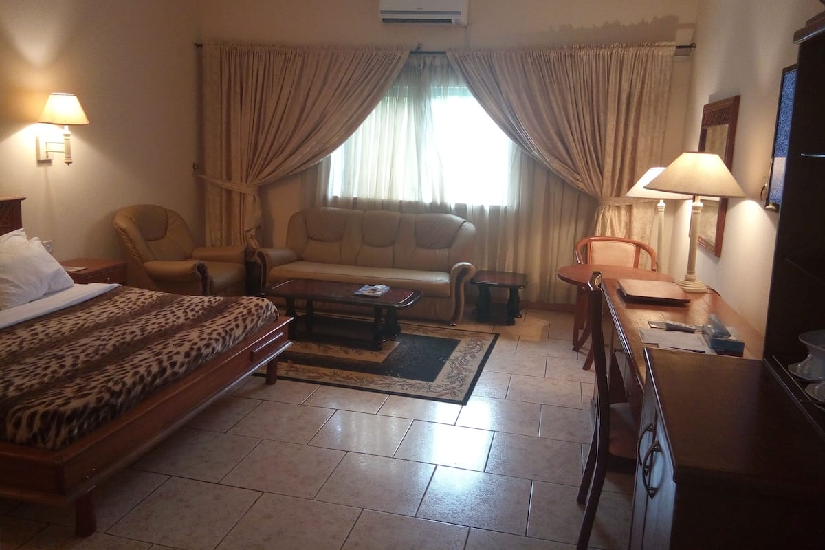 A hotel room with a bed, a sofa, a chair, a small table, and an air conditioning unit.