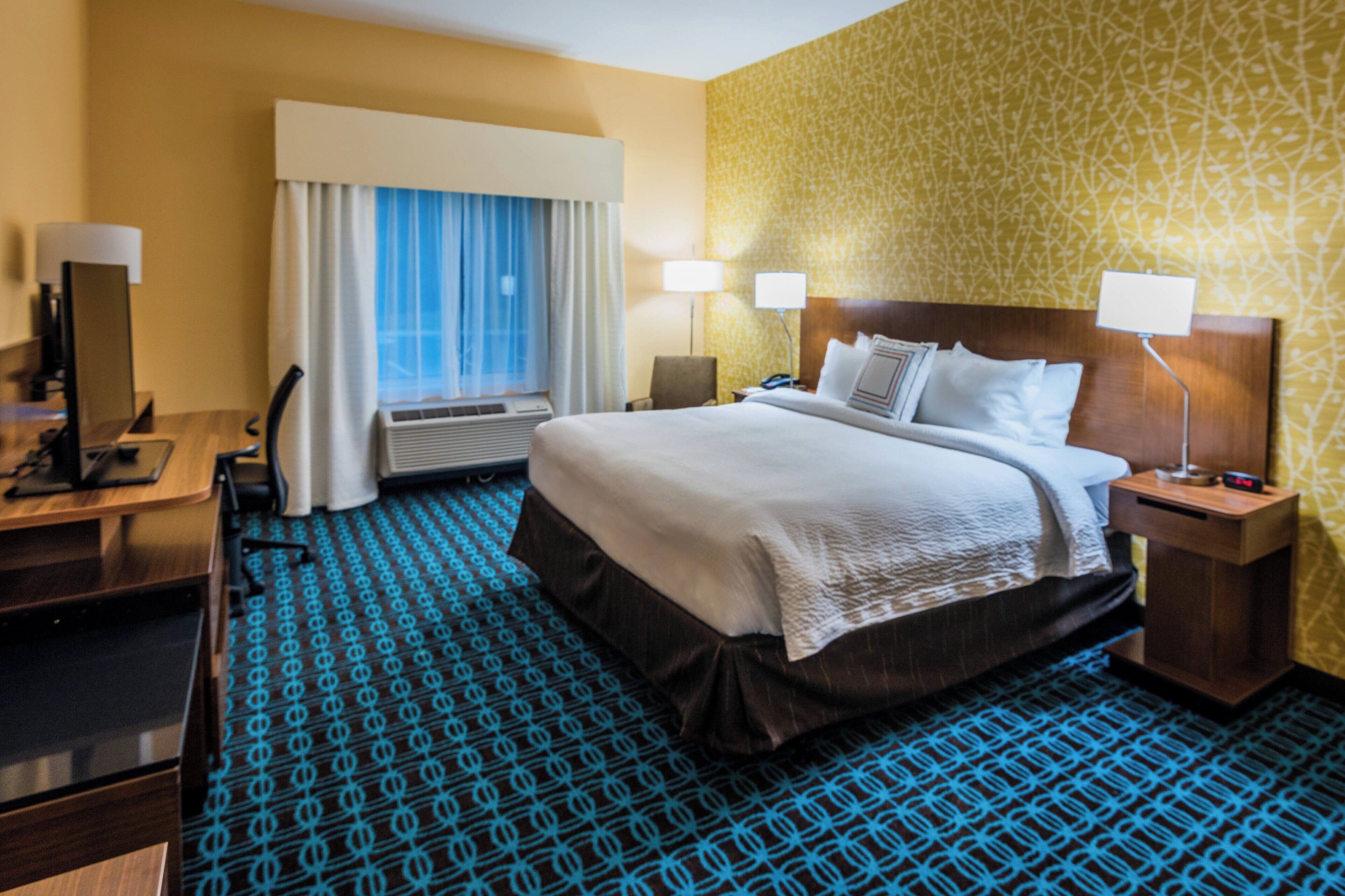 A hotel room with a large bed, two bedside tables with lamps, a desk with a chair, and a window with curtains.