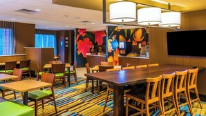 A hotel dining area with wooden tables and chairs, a colorful striped carpet, and a large flat-screen TV on the wall. - Fairfield Inn & Suites by Marriott Atlanta Fairburn (Fairburn)