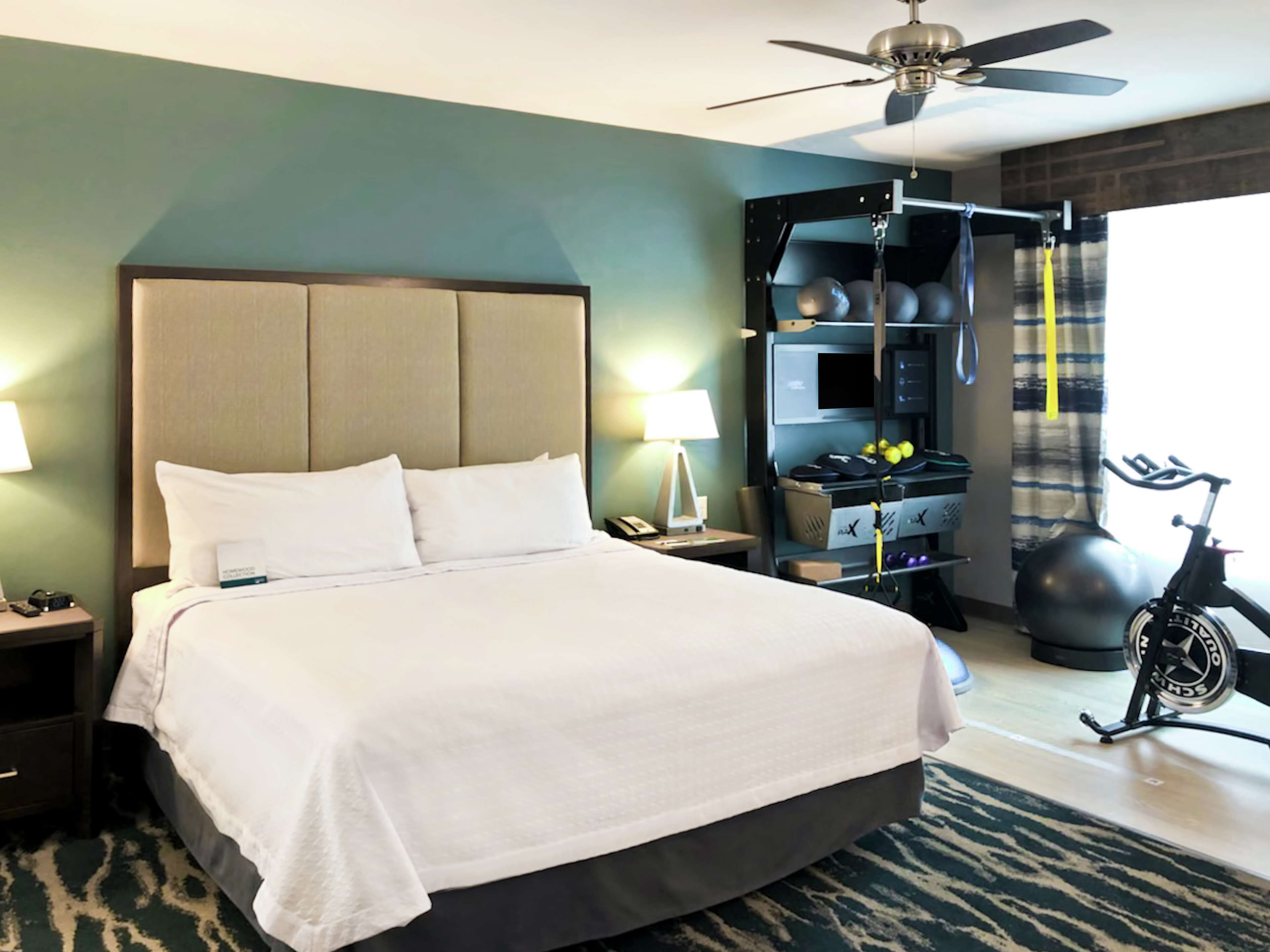 Una habitación de hotel con una cama grande, un ventilador de techo, un espacio para ejercitarse con equipamiento y vistas al exterior.