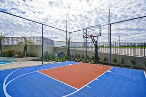 Ein Basketballplatz im Freien mit einer blau-orangenen Spielfläche, umgeben von einem Metallzaun und einem Schwimmbecken.