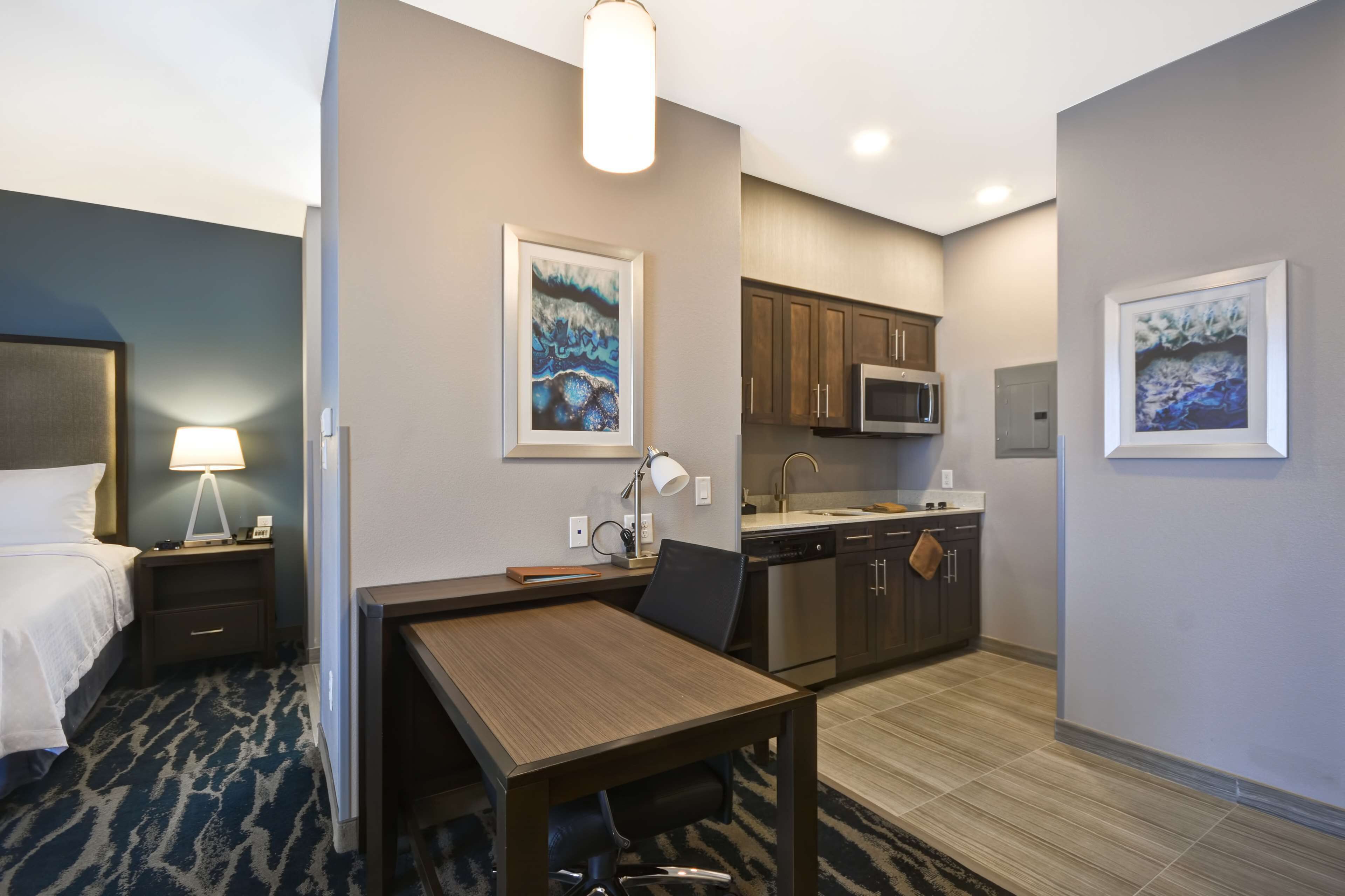 A hotel room with a bed, a desk, a chair, a lamp, a microwave, a sink, and cabinets.