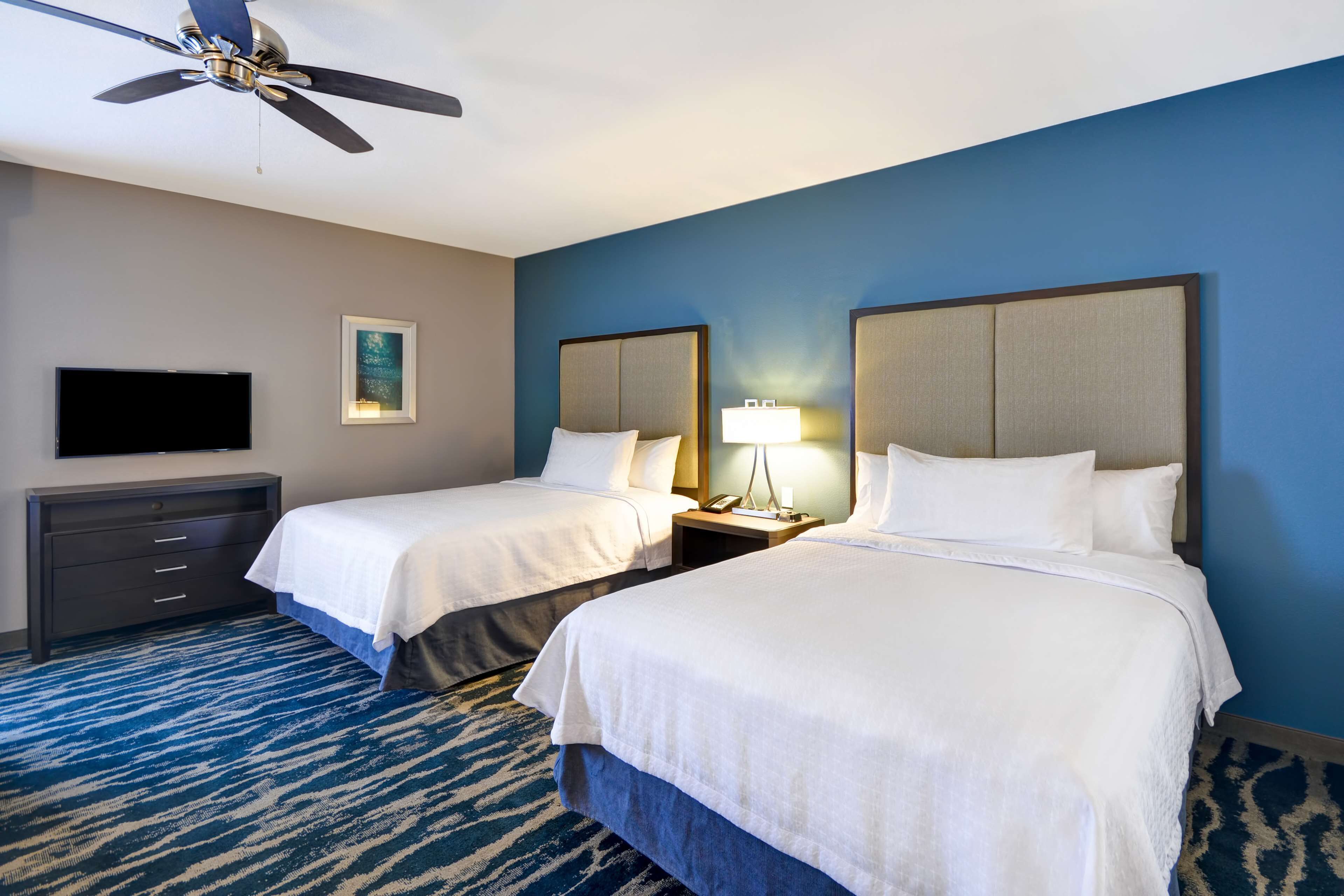 A hotel room with two beds, a ceiling fan, a flat-screen TV, a desk with drawers, a lamp, and a framed picture on the wall.