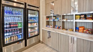 A self-service snack and beverage area with refrigerated drink dispensers and shelves stocked with snacks and beverages. - Homewood Suites by Hilton Conroe (Conroe)