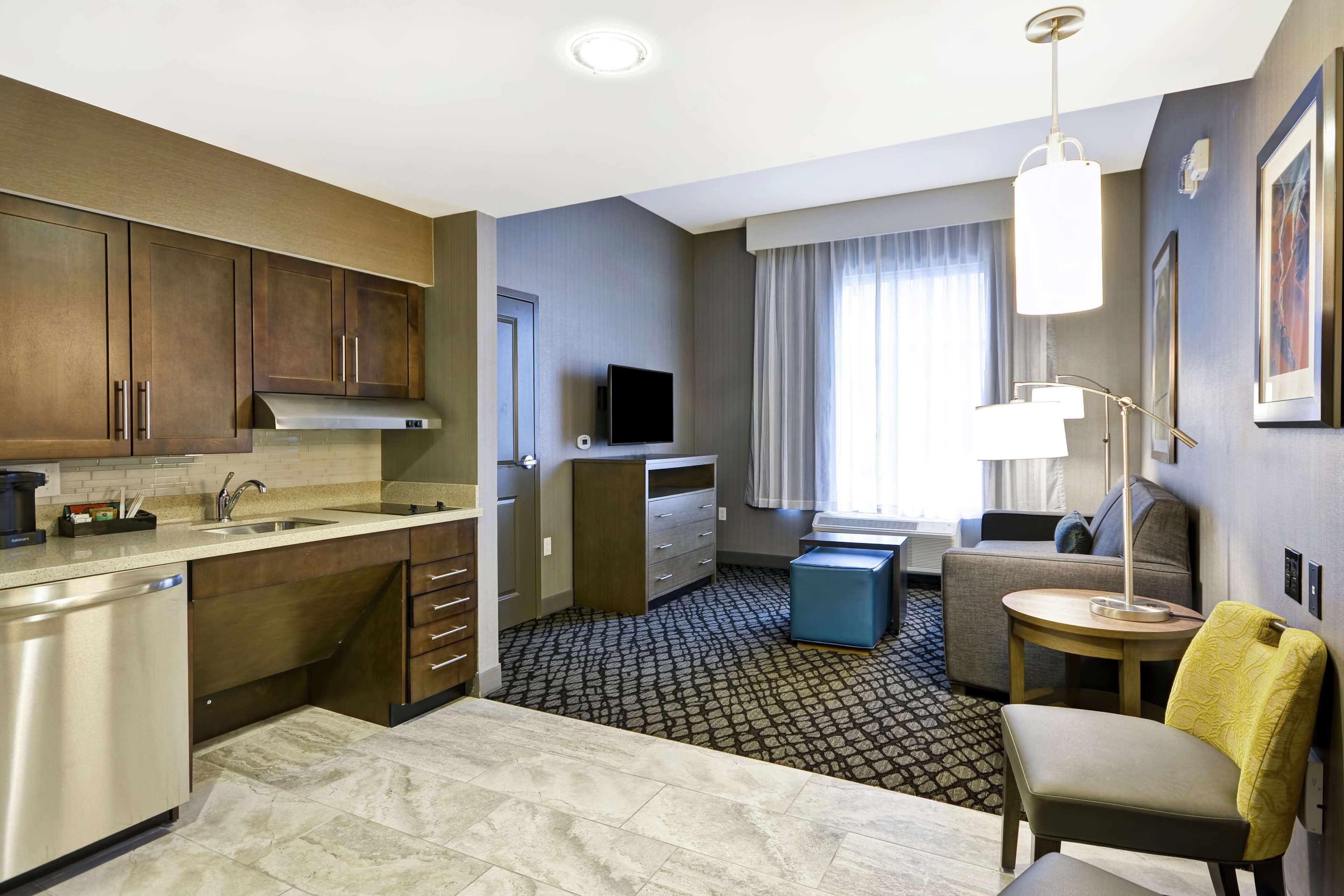 A compact hotel room with a kitchenette, a seating area with a sofa and armchair, a small table, and a TV.