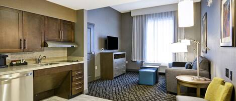 A compact hotel room with a kitchenette, a seating area with a sofa and armchair, a small table, and a TV.