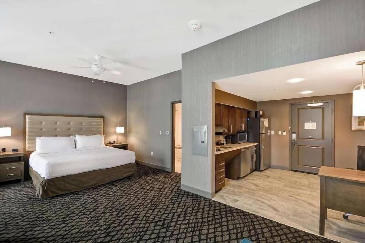 A hotel room with a bed, a desk, a kitchenette, and a ceiling fan.