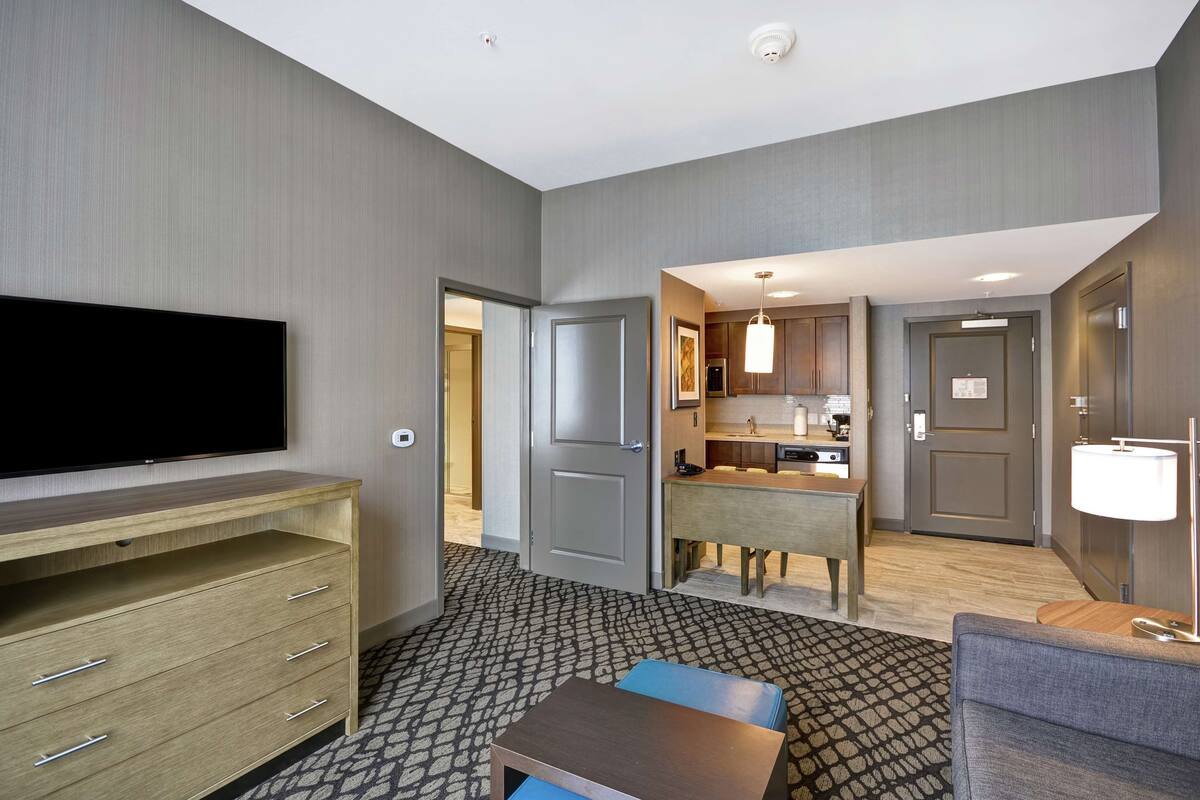 A hotel room with a flat-screen TV, a wooden dresser, a patterned carpet, a small table, a sofa, and a kitchenette.