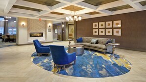A spacious lobby with a fireplace, blue armchairs, a round coffee table, and framed artwork on the walls. - Homewood Suites by Hilton Detroit (Warren)
