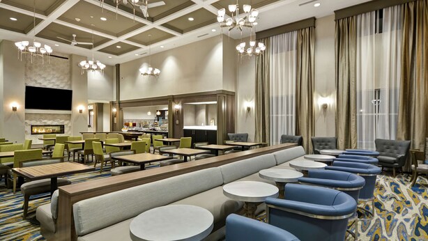 A hotel lobby with a long bench, round tables, and blue chairs. There are chandeliers, a fireplace, and a large window with curtains. - Homewood Suites by Hilton Detroit (Warren)