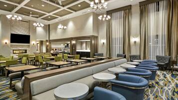 En hotellobby med en lång bänk, runda bord och blå stolar. Här finns kristallkronor, en öppen spis och ett stort fönster med gardiner.