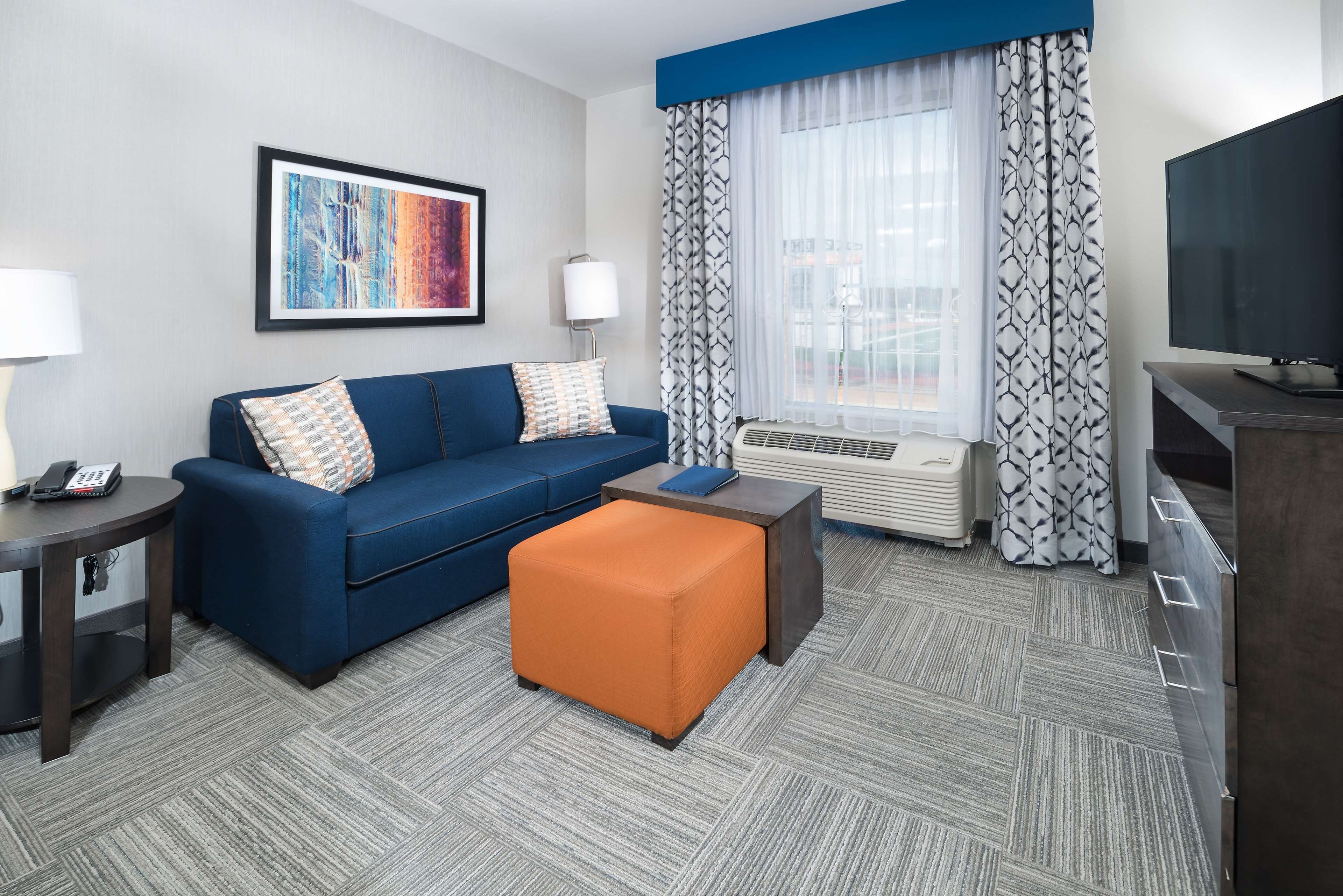 A hotel room with a blue sofa, an orange ottoman, a flat-screen TV, and a large window with patterned curtains.