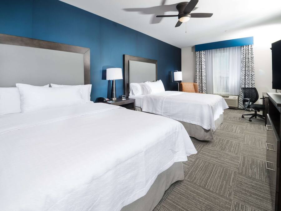 A hotel room with two beds, a desk, a chair, a television, and a ceiling fan.