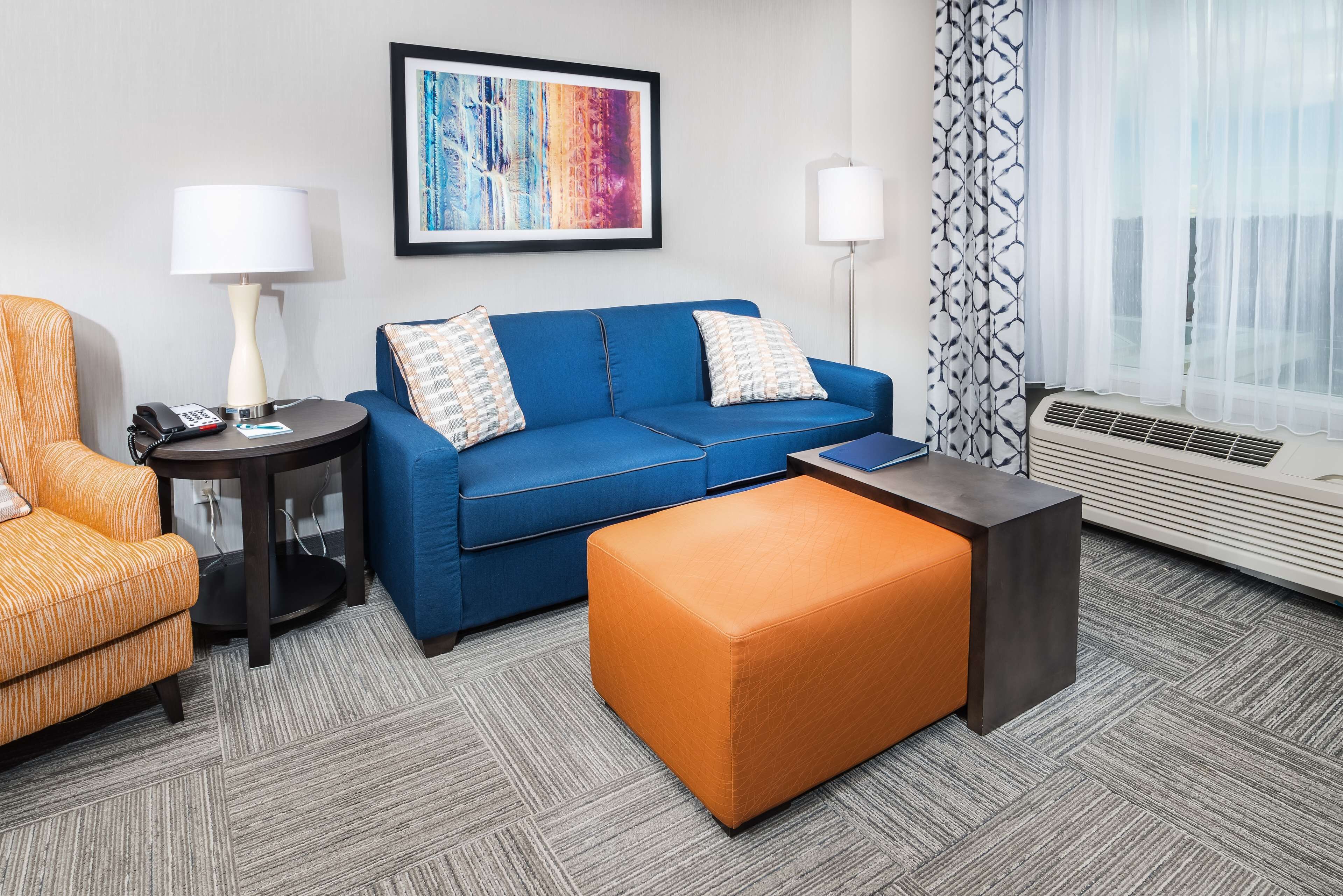 Une chambre d’hôtel avec un canapé bleu, un fauteuil orange, une petite table ronde avec une lampe, un tableau abstrait encadré et une grande fenêtre avec des rideaux légers.
