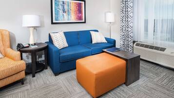 Une chambre d’hôtel avec un canapé bleu, un fauteuil orange, une petite table ronde avec une lampe, un tableau abstrait encadré et une grande fenêtre avec des rideaux légers.