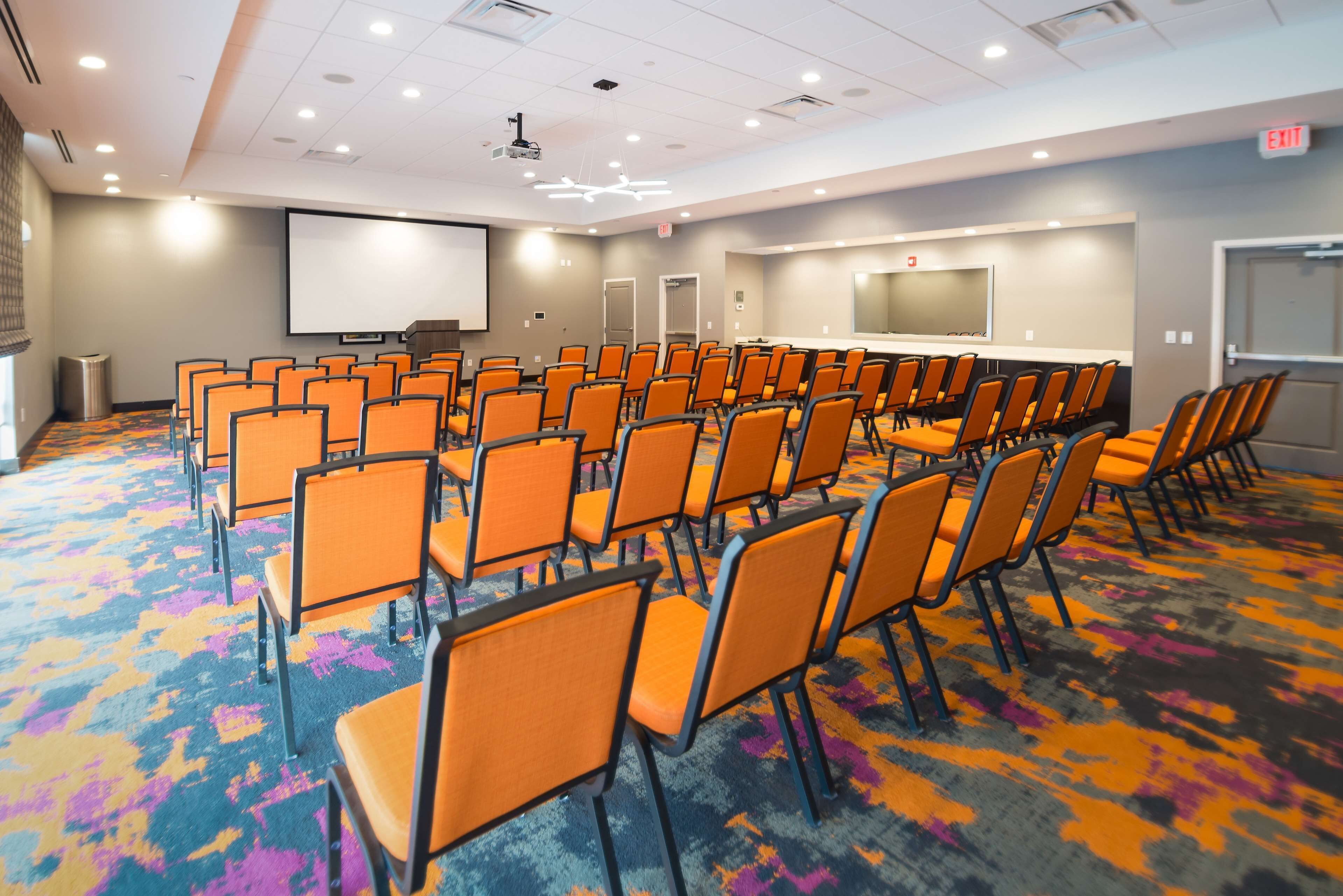 Een vergaderzaal met rijen oranje stoelen, een scherm voor een projector en een vloerbedekking.