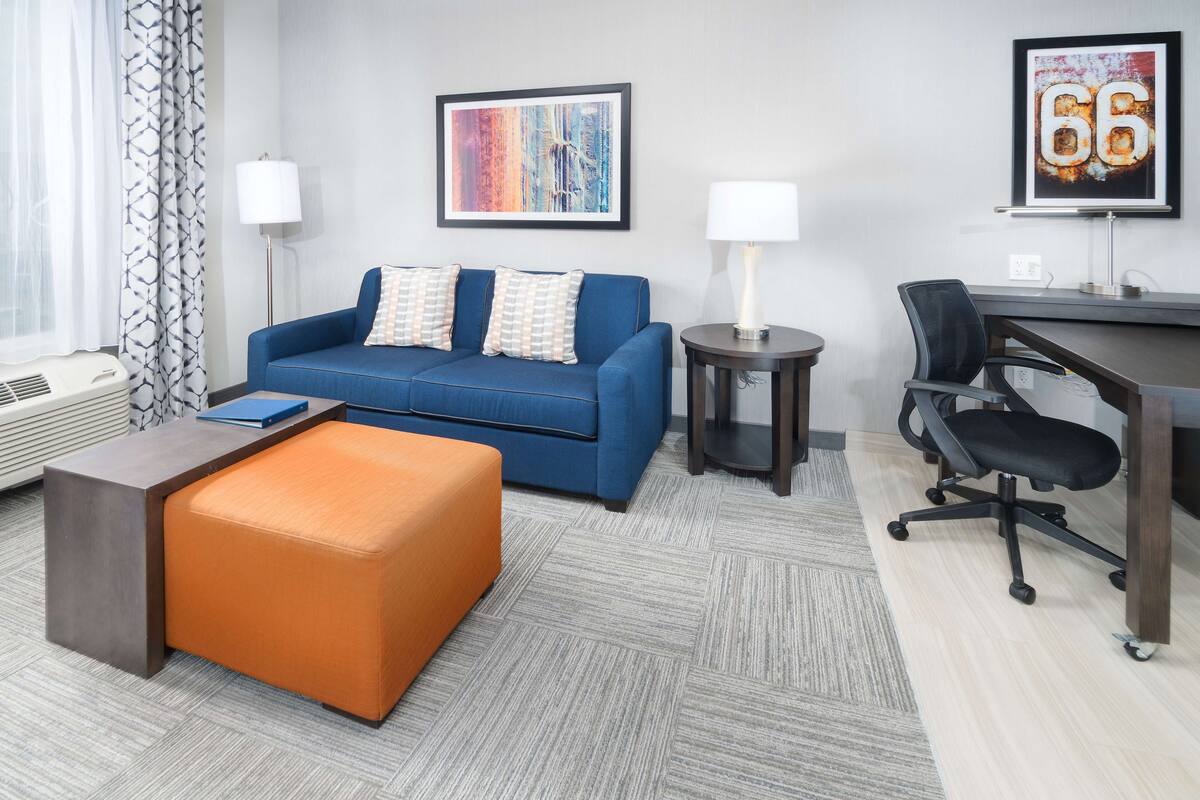 Une chambre d’hôtel moderne avec un canapé bleu, un pouf orange, un bureau avec une chaise et deux lampes.