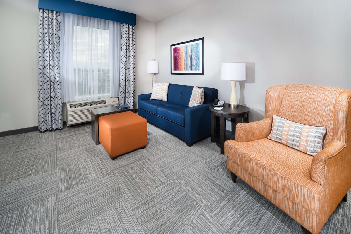 Une chambre d’hôtel avec un canapé bleu, un fauteuil orange, un petit pouf orange, une table d’appoint avec une lampe et une grande fenêtre ornée de rideaux l égers.