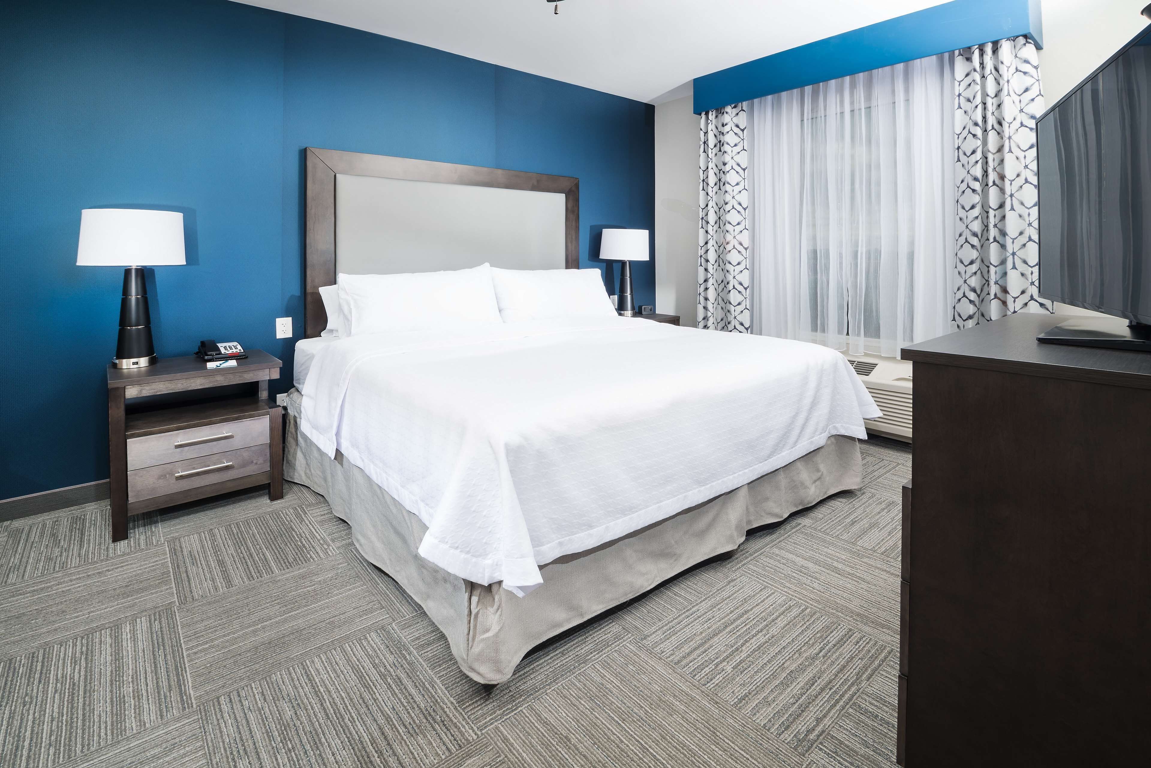 A hotel room with a large bed, two bedside tables with lamps, a dresser, and a window with patterned curtains.
