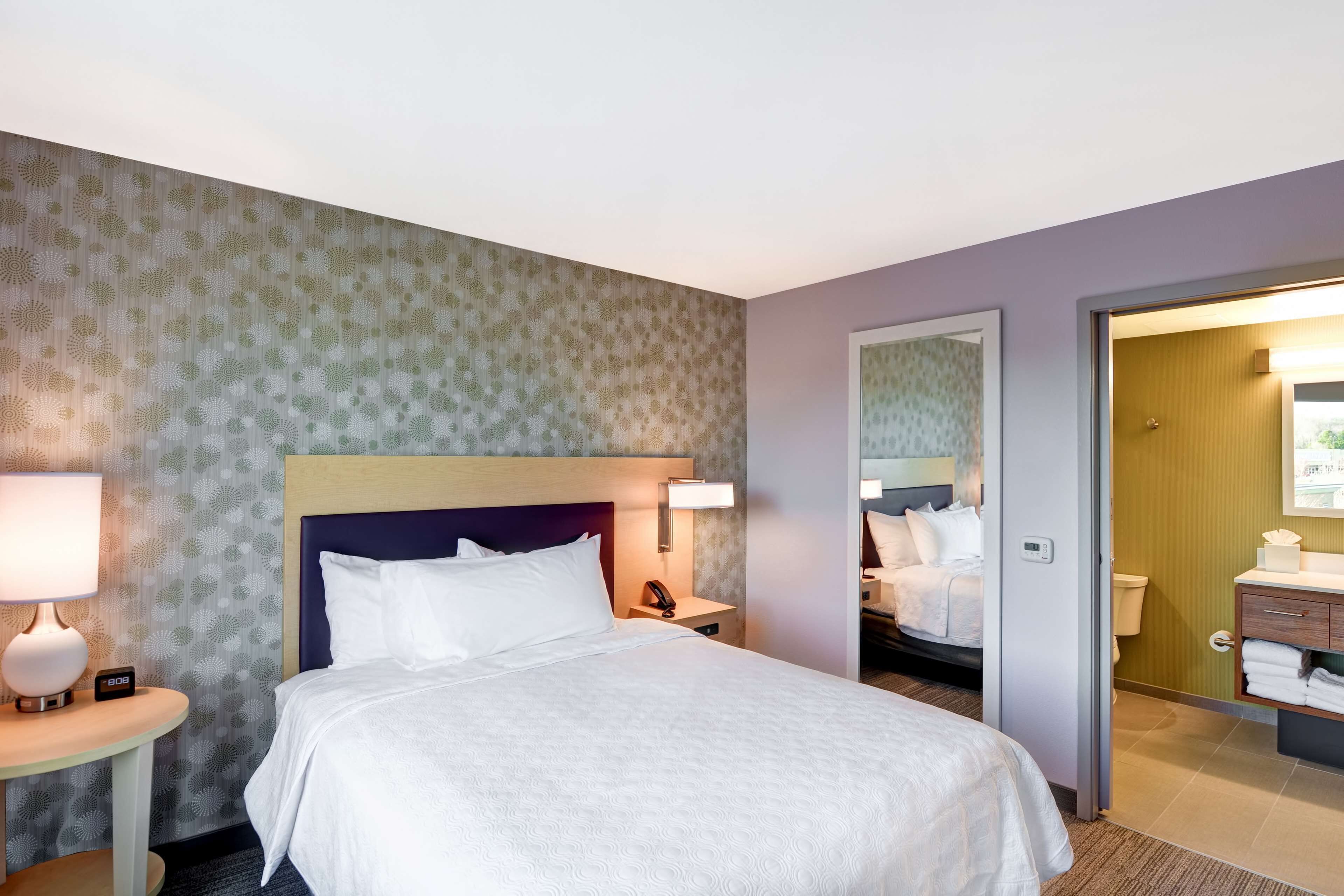A hotel room with a neatly made bed, a bedside table with a lamp, a nightstand with a phone, and a bathroom visible through an open door.