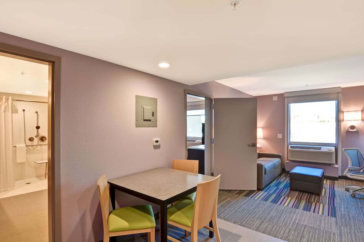 A hotel room with a dining area, a shower, a bed, and a window with blinds.