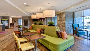 Un vestíbulo moderno con un sofá verde, mesas de madera y un sillón azul. Hay plantas en macetas, un mostrador de recepción y una pared de piedra.