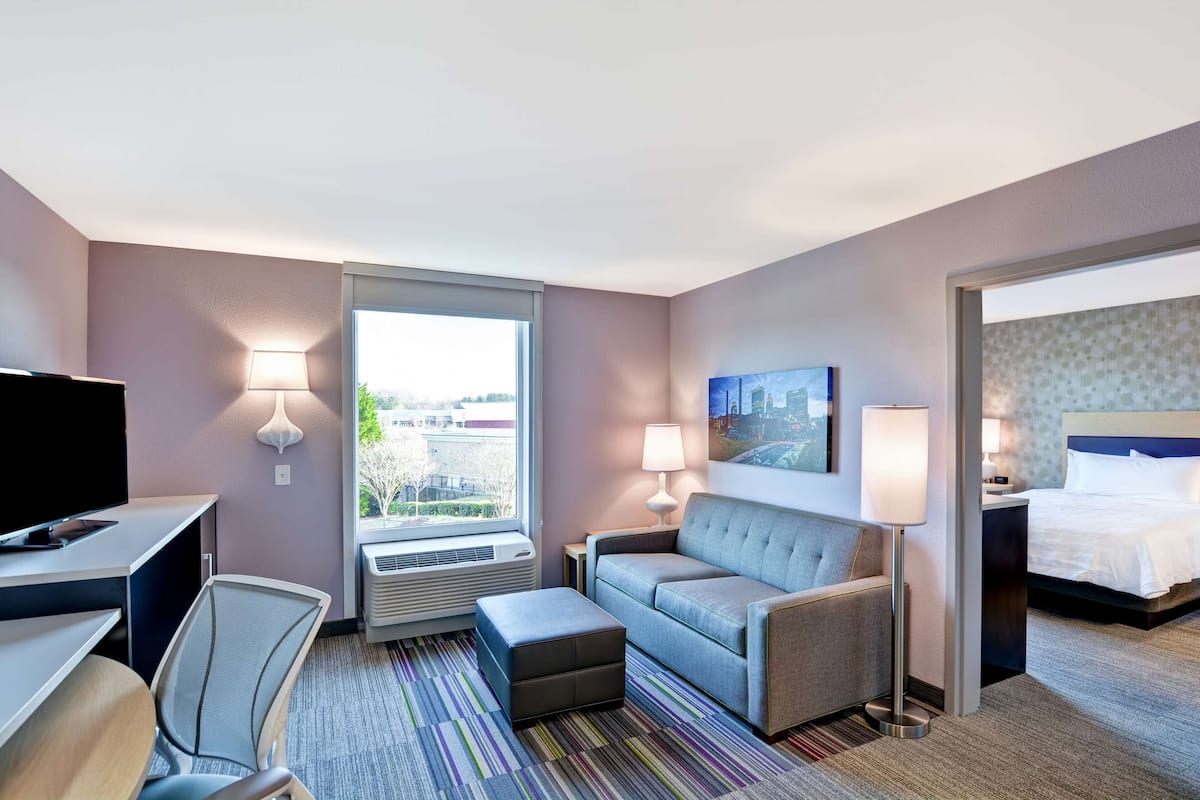 A hotel room with a bed, a sofa, a desk with a TV, a chair, and a window with a view of buildings.