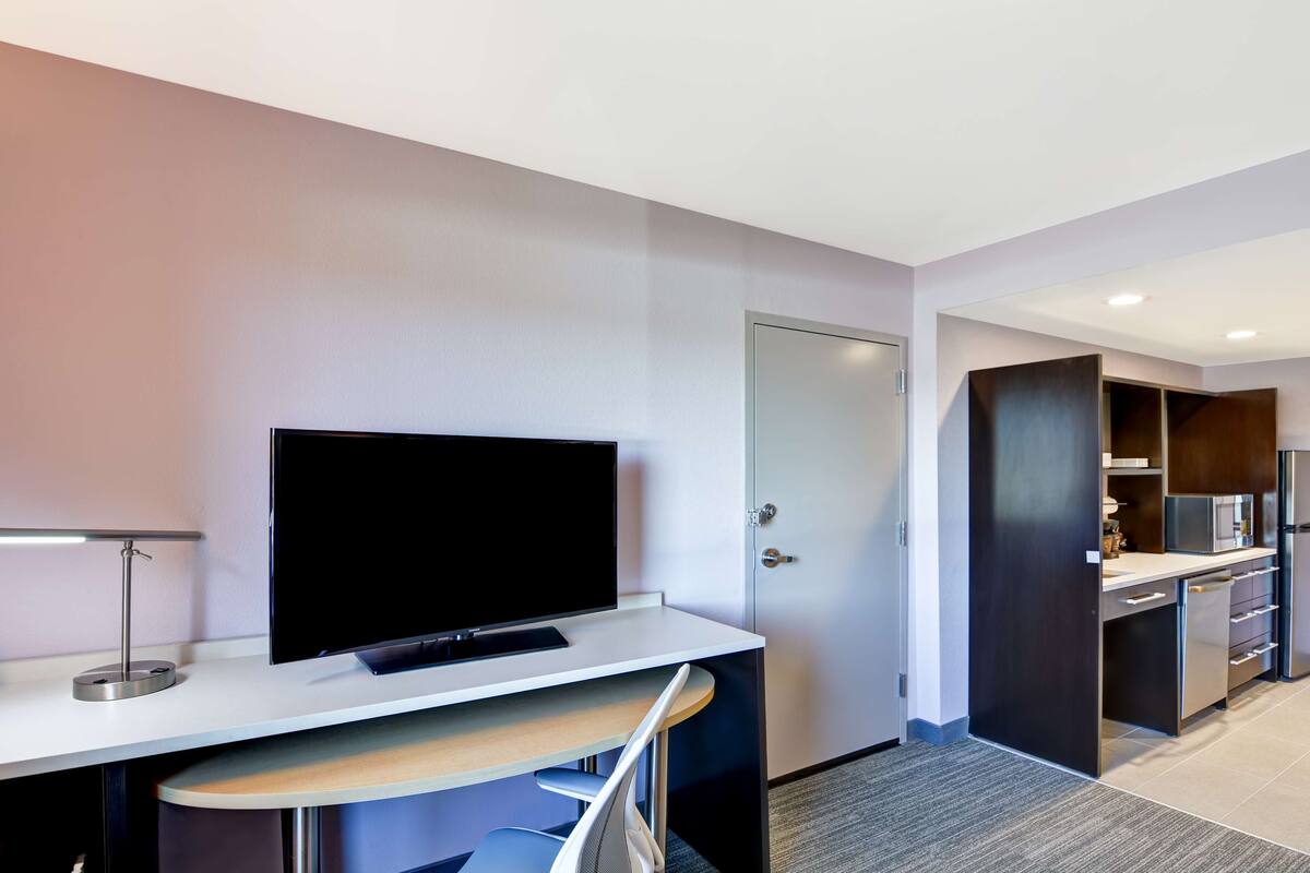A modern hotel room with a flat-screen TV on a white desk, a built-in kitchen with a microwave, and a door leading to a closet.