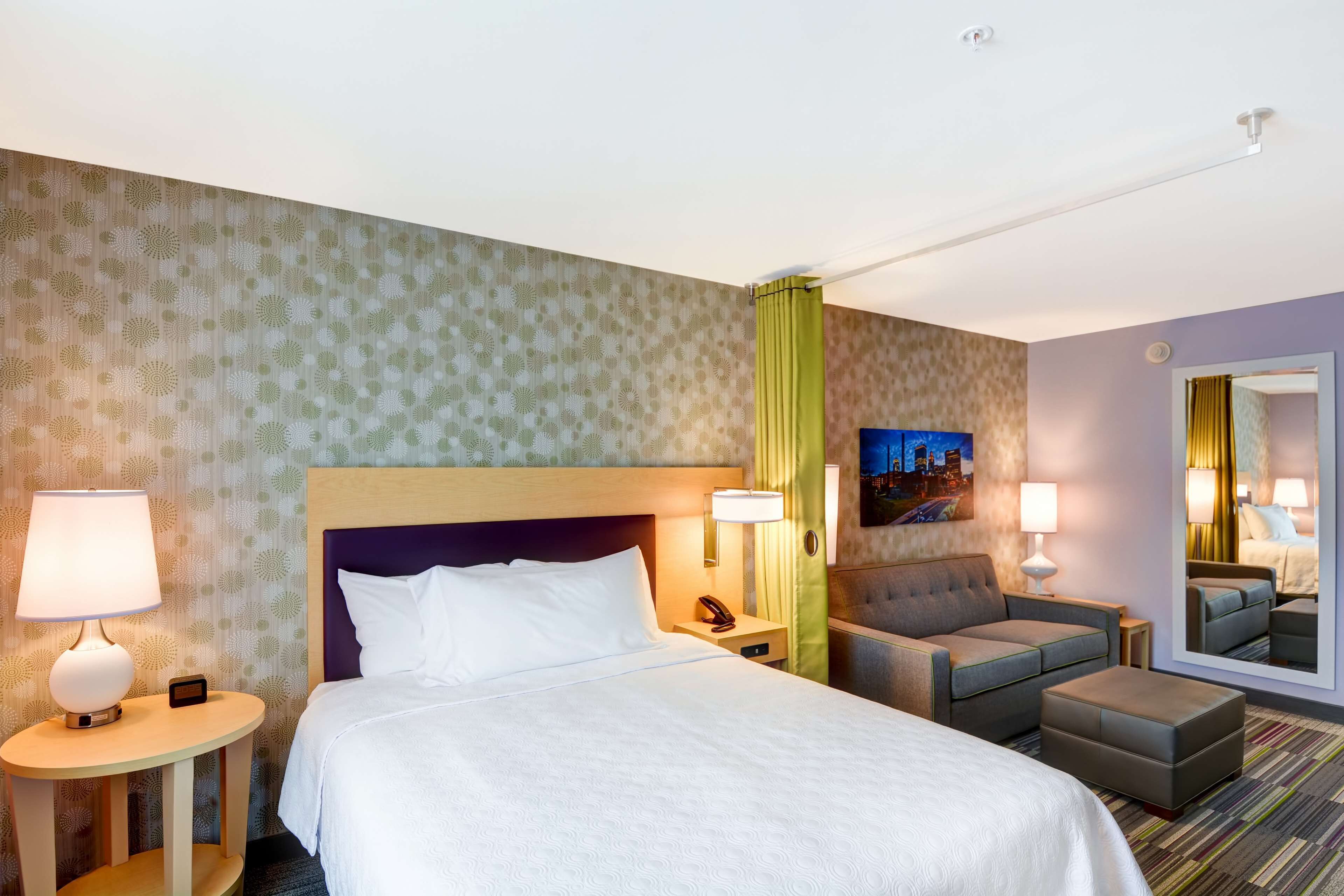 A hotel room with a large bed, a sofa, a small table, and a mirror.