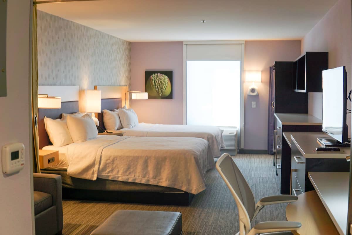 A hotel room with two beds, a desk, a chair, a TV, and a window.