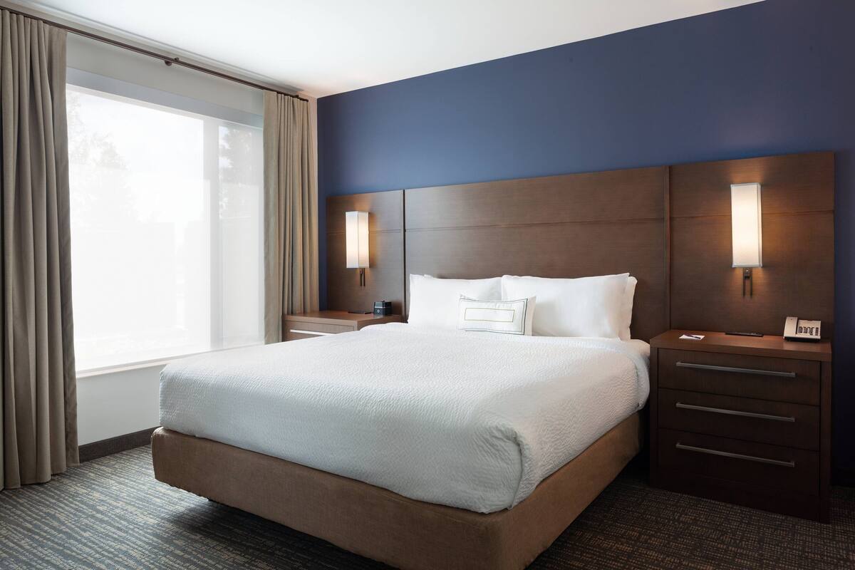 Habitación de hotel con una cama grande, una mesita de noche con lámpara y vistas al exterior a través de una ventana.
