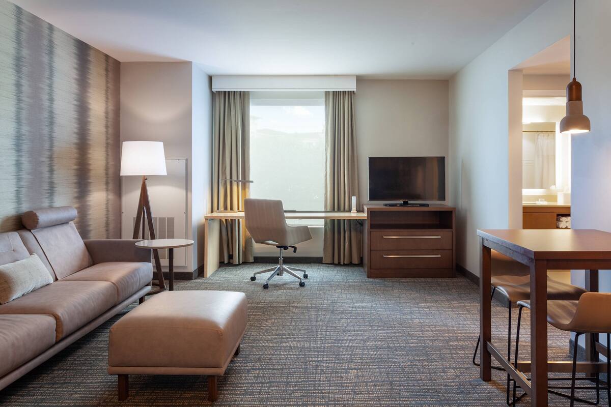 Habitación de hotel con sofá, reposapiés, escritorio, silla, televisión y vistas al exterior.