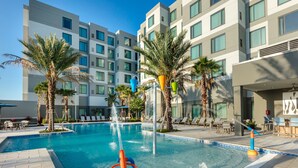 A modern hotel pool with a water feature, surrounded by palm trees and lounge chairs.