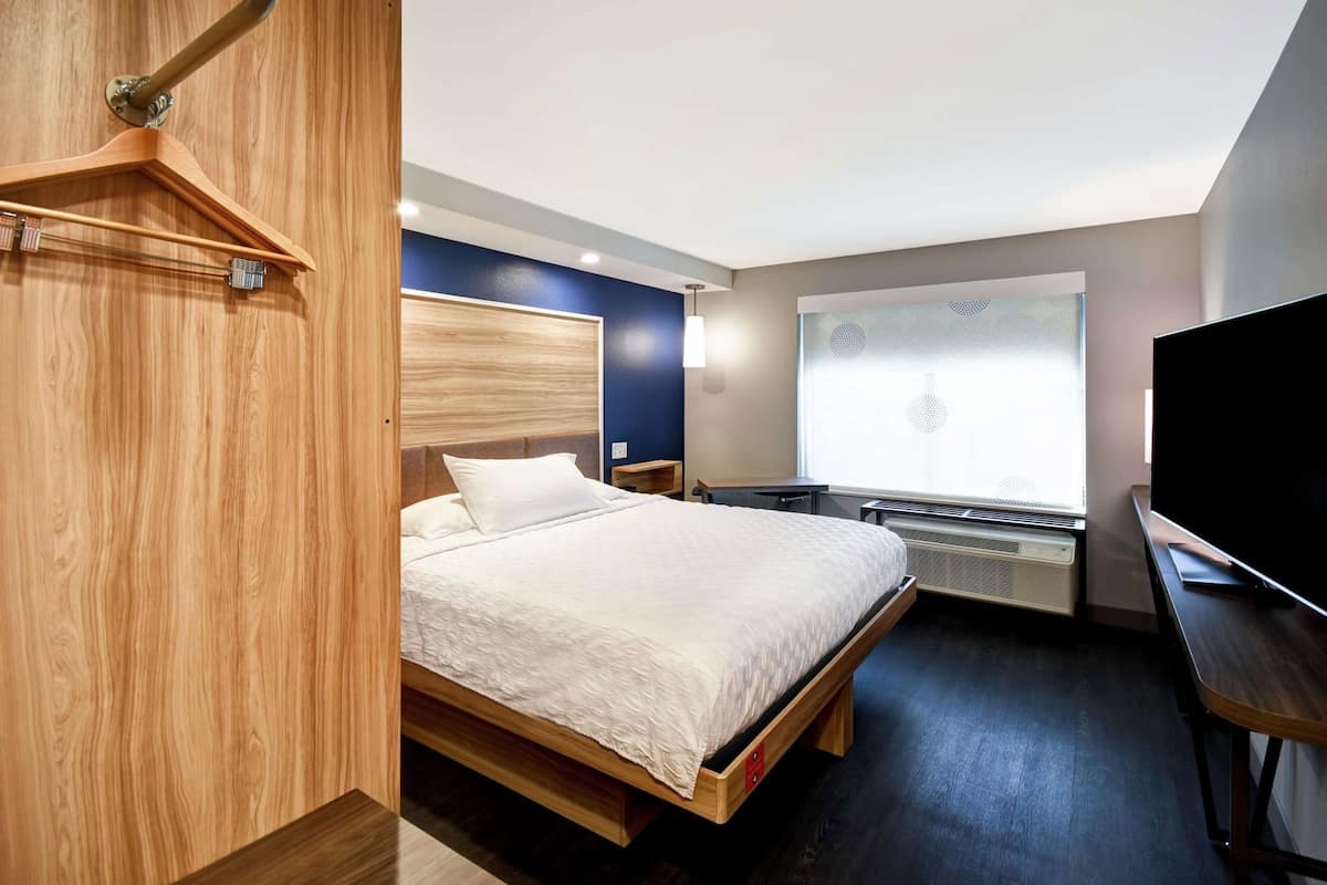 A modern hotel room with a wooden bed, a flat-screen TV, a desk, and a window with blinds.