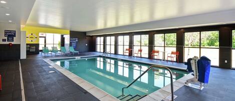 Una piscina interior con suelo de baldosas, una zona junto a la piscina con sillas y vistas al exterior a través de amplias ventanas.