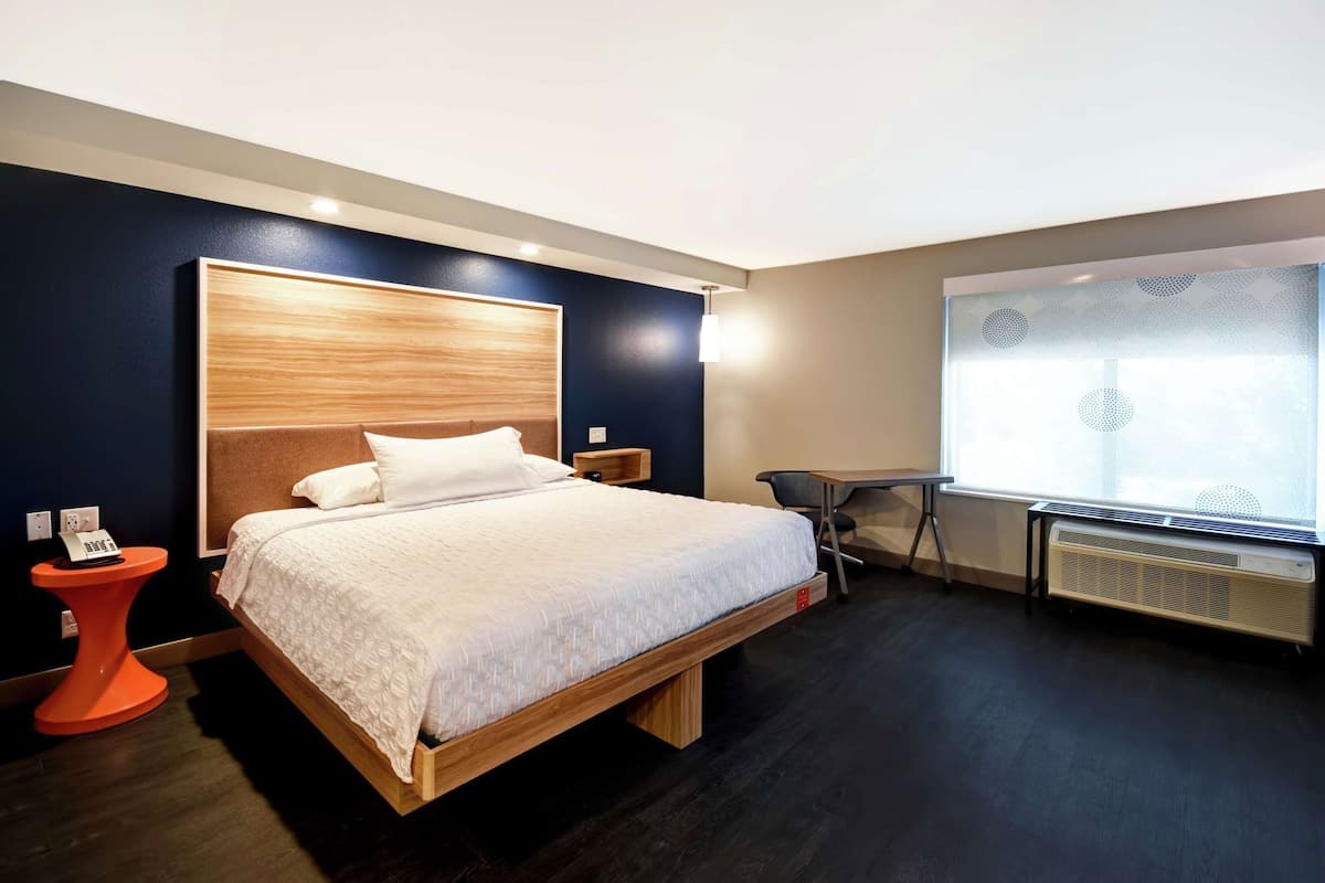 A modern hotel room with a wooden bed, a desk, a chair, a telephone, and a window with a view.