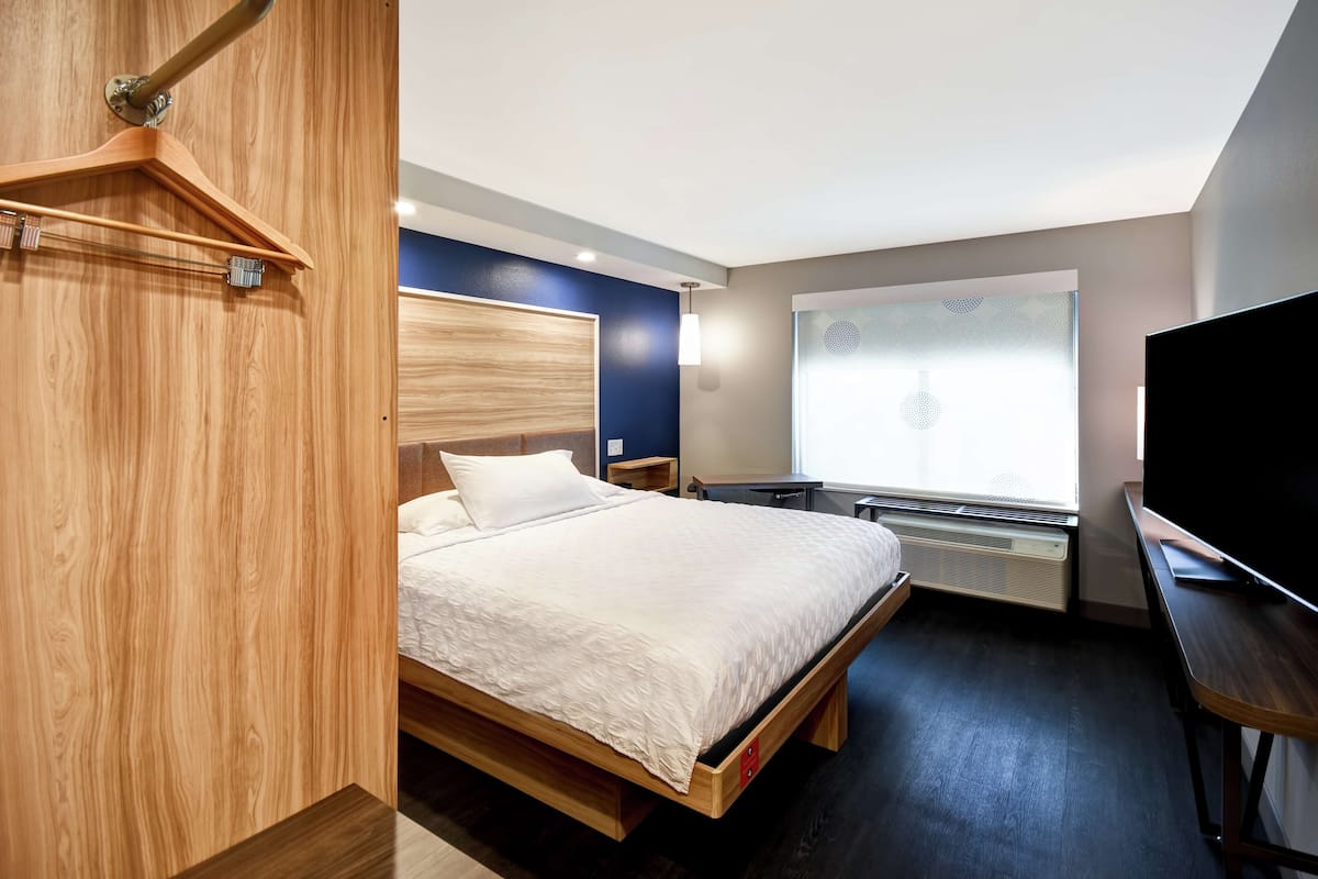 A modern hotel room with a wooden bed frame, a flat-screen TV, a desk, and a window with a view.